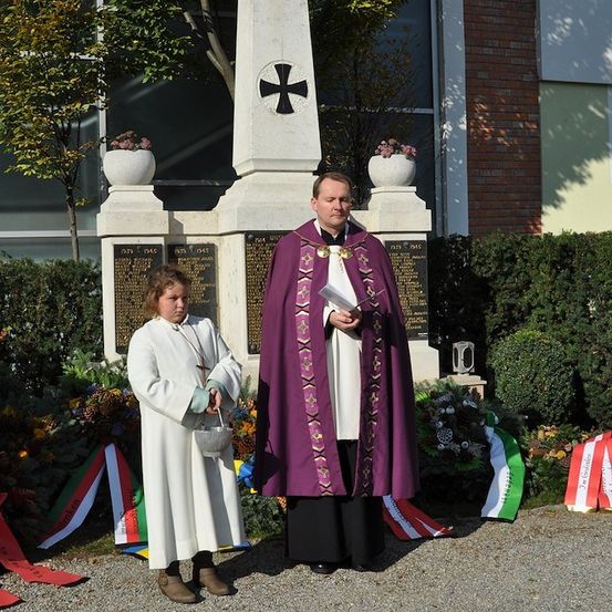Ein Priester in einem lila Gewand steht neben einem jungen Mädchen in einem weißen Gewand, beide halten einen Korb, vor einem Denkmal mit einem Kreuz und Tafeln, umgeben von Blumen und Flaggen.