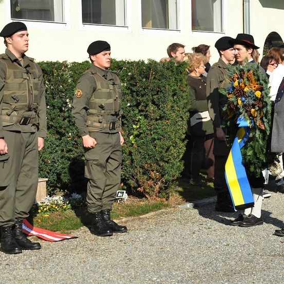 Eine Gruppe von Soldaten in Uniform steht vor einem Gebäude, wobei ein Mann eine Girlande hält.