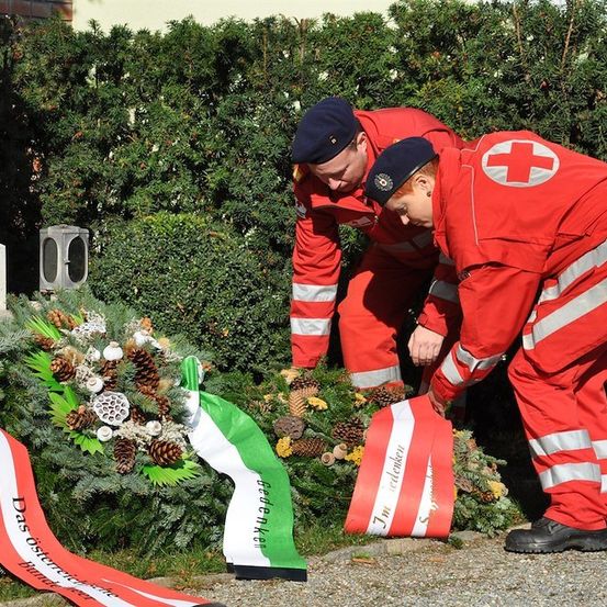 Zwei Personen in roten Uniformen mit weißen Streifen und roten Mützen platzieren einen Kranz mit Tannenzapfen und Bändern auf einem Gedenkstein.