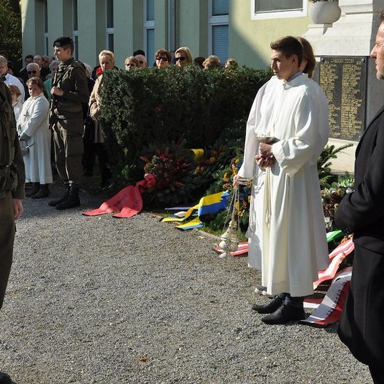 Eine feierliche Zeremonie mit einem Priester und Militärangehörigen vor einem Denkmal mit Blumen und Flaggen.