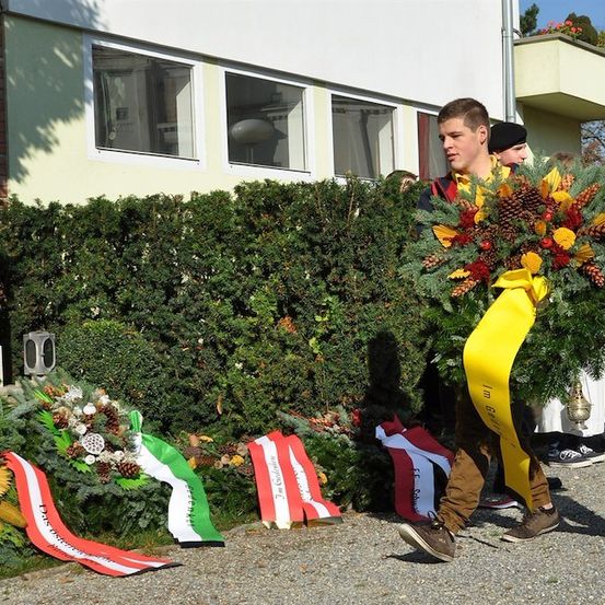 Ein junger Mann trägt einen Kranz aus Blumen und Blättern und geht an einem Denkmal mit Fahnen und einem Gebäude im Hintergrund vorbei.