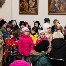Eine Gruppe von Kindern in Winterkleidung steht in einem Raum mit zwei Gemälden an der Wand. Einige tragen Mützen und Schals. Eine Frau mit Brille sitzt vor ihnen.