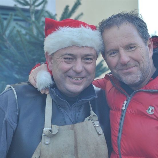 Zwei Männer lächeln und posieren für ein Foto vor einem Weihnachtsbaum. Einer trägt einen Weihnachtsmannhut und eine Schürze.
