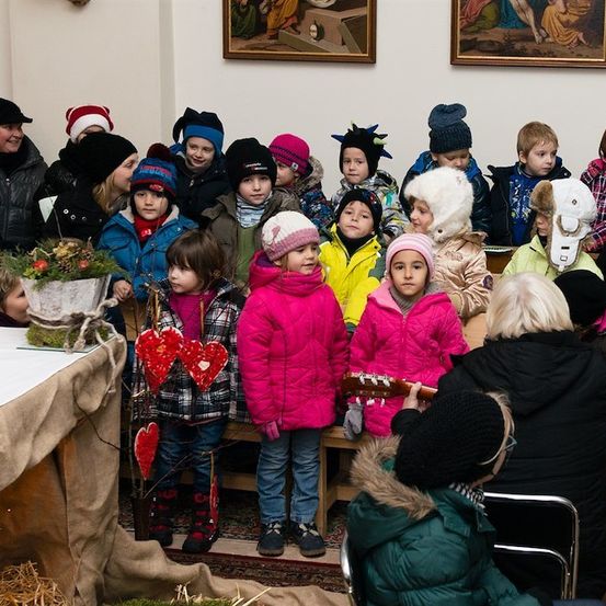 Eine Gruppe von Kindern in Winterkleidung steht in einem Raum, einige tragen Mützen und Handschuhe. Eine Frau spielt vor ihnen eine Gitarre. Ein Tisch mit einem Topf steht links.
