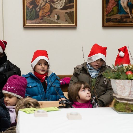 Eine Gruppe von Kindern in Winterkleidung und Weihnachtsmützen versammelt sich um einen Tisch mit einer Pflanze und Kerzen. Sie befinden sich in einem Raum mit Gemälden an der Wand.