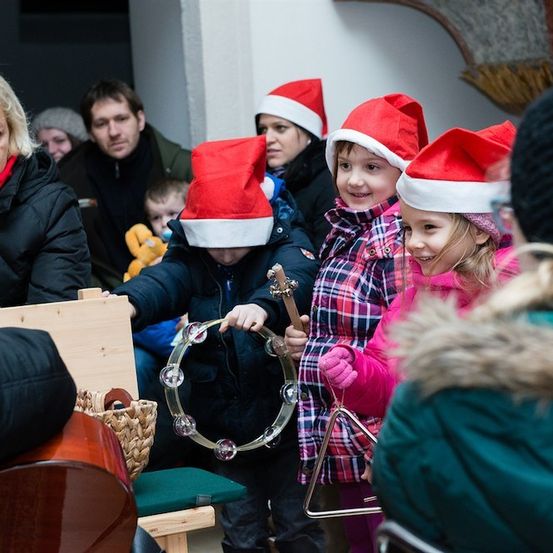 Eine Gruppe von Kindern mit Weihnachtsmützen spielt Musikinstrumente, darunter eine Schelle und ein Cello, in einem Raum, in dem Erwachsene zusehen.