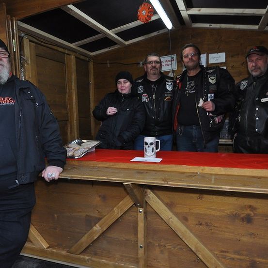 Fünf Personen, vier Männer und eine Frau, stehen hinter einem Holztresen. Sie tragen alle Lederjacken. Der Mann auf der linken Seite trägt einen schwarzen Hut und eine Brille. Ein Becher mit Totenkopf-Design steht auf dem Tresen.