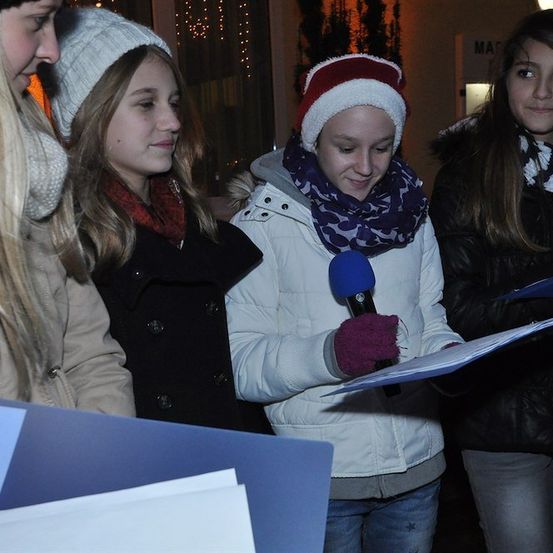 Eine Gruppe junger Mädchen in Winterkleidung, eine hält ein Mikrofon und liest von einem Stück Papier. Sie stehen vor einem Gebäude mit dekorativen Lichtern.