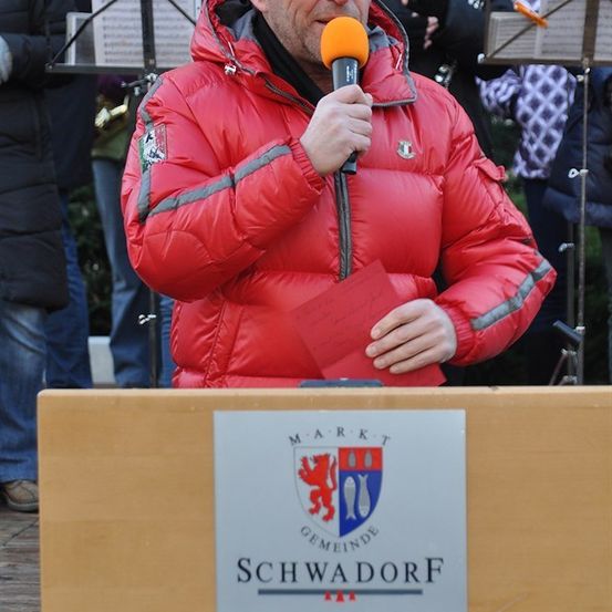 Ein Mann in einer roten Daunenjacke steht an einem Podium, hält ein Mikrofon und ein rotes Papier in der Hand, mit einem Schild, das 'Marktgemeinde Schwadorf' vor ihm steht. Dahinter halten Musiker Notenständer.