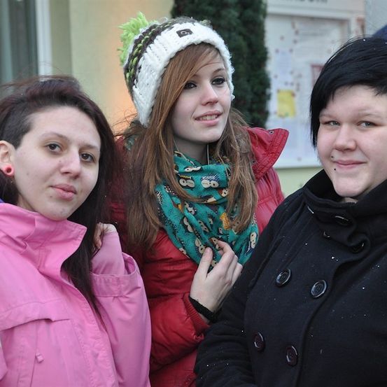 Drei Frauen stehen zusammen, eine trägt eine rosa Jacke, eine andere eine rote Jacke mit einem grünen Schal und die dritte eine schwarze Jacke. Sie lächeln und scheinen für ein Foto zu posieren.