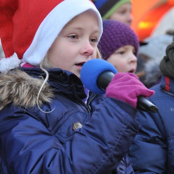 Ein junges Mädchen mit Weihnachtsmütze und Handschuhen hält ein Mikrofon und singt in einer Menschenmenge von Kindern.