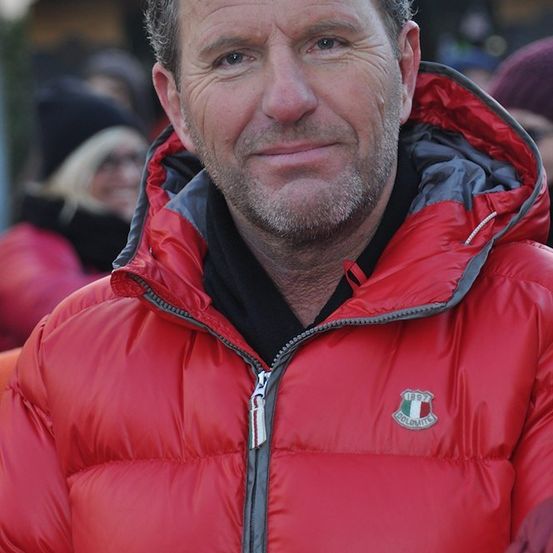 Ein Mann mit Bart und Schnurrbart, der eine rote Jacke mit einem Logo auf der linken Seite seiner Brust trägt. Er lächelt und schaut in die Kamera. Hinter ihm sind verschwommene Menschen, möglicherweise bei einer Veranstaltung.