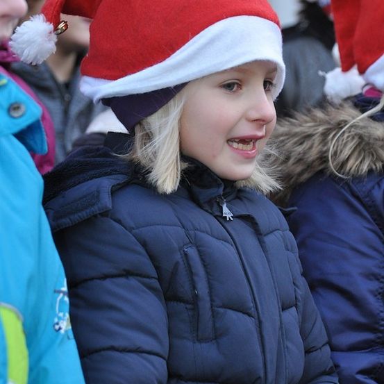 Ein junges Mädchen mit blonden Haaren, das eine rote und weiße Weihnachtsmütze und eine blaue Jacke trägt, lächelt in einer Menschenmenge.