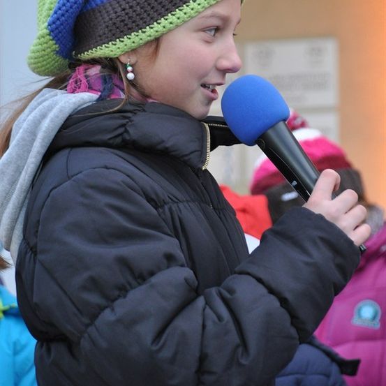 Ein junges Mädchen mit einer bunten Mütze und einer schwarzen gefütterten Jacke hält ein blaues Mikrofon und scheint in dieses zu sprechen.