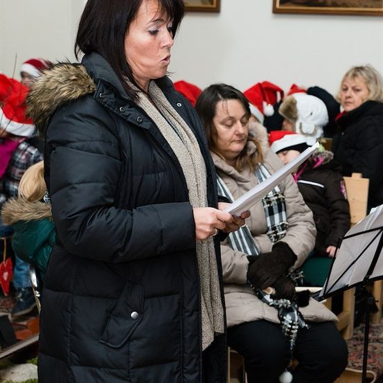 Eine Frau in einem schwarzen Mantel mit Pelzkragen steht und liest ein Stück Papier, während andere hinter ihr sitzen und Weihnachtsmützen tragen.
