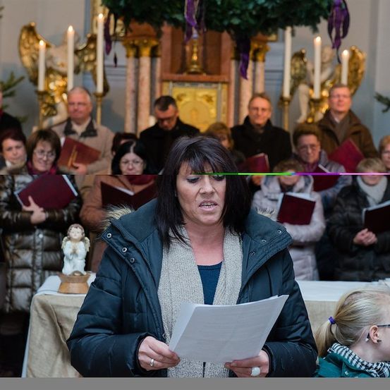 Eine Frau liest ein Papier vor einer Gruppe von Menschen in einer Kirche. Die Gruppe hält Bücher und scheint zu singen.