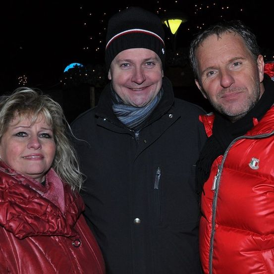 Drei Menschen stehen eng beieinander und lächeln für ein Foto. Der Mann in der Mitte trägt eine schwarze Jacke und eine Mütze. Die Frau auf der linken Seite trägt eine rote Jacke. Der Mann auf der rechten Seite trägt eine rote Daunenjacke. Sie befinden sich in einem dunklen Bereich mit einigen Lichtern im Hintergrund.