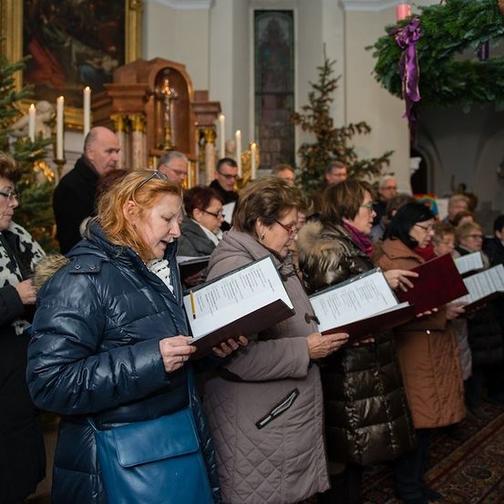 Eine Gruppe von Menschen in Winterkleidung versammelt sich in einer Kirche und singt aus Gesangbüchern. Die Kirche ist mit Weihnachtsbäumen und Kerzen geschmückt.