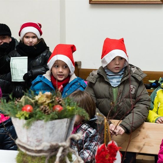 Eine Gruppe von Kindern in Winterkleidung und Weihnachtsmützen sitzt in einer Kirche, einige halten Papiere und andere schauen auf eine dekorierte Pflanze.