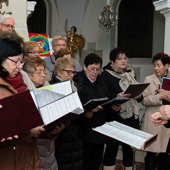 Eine Gruppe älterer Frauen und Männer versammelt sich in einer Kirche, liest aus Gesangbüchern und singt. Ein Mann steht in der Nähe, möglicherweise als Chorleiter.
