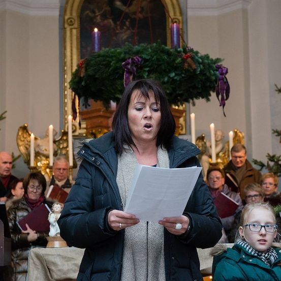 Eine Frau steht vor einem weihnachtlich geschmückten Altar und singt, während sie ein Stück Papier in der Hand hält. Hinter ihr singt eine Gruppe von Menschen, einige halten Bücher. Der Altar ist mit Kerzen und einem Kranz geschmückt.