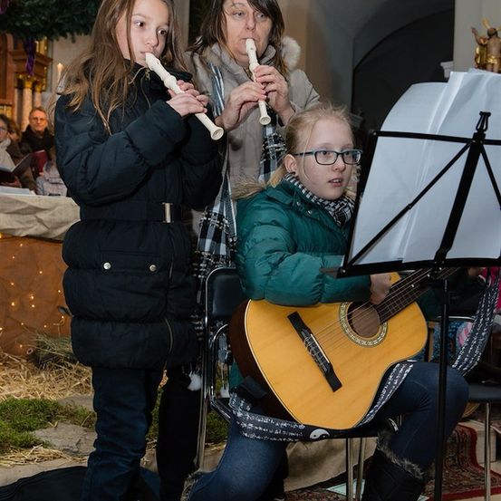 Drei Personen spielen in einer Kirche Musikinstrumente. Ein Mädchen links spielt eine Blockflöte, ein anderes Mädchen in der Mitte spielt Gitarre und eine Frau steht hinter ihnen und spielt ebenfalls eine Blockflöte.