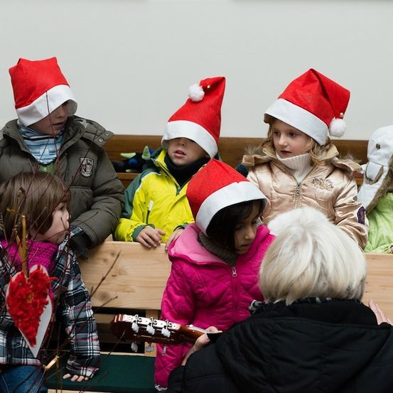 Eine Gruppe von Kindern mit Weihnachtsmützen sitzt auf einer Bank, eines von ihnen hält eine Gitarre, während eine erwachsene Frau sie ansieht.