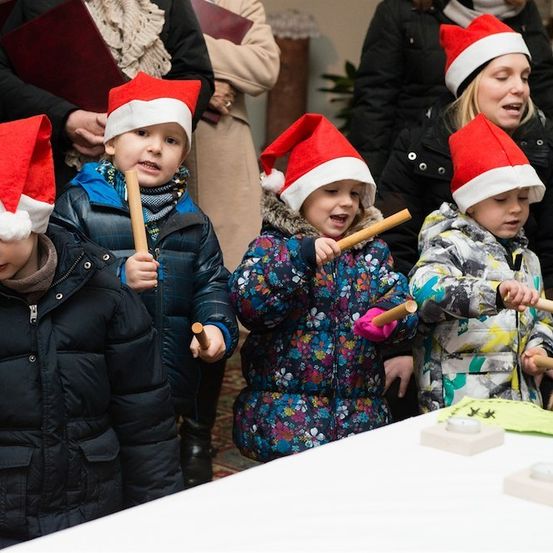Eine Gruppe von Kindern, die in Winterkleidung und Weihnachtsmützen gekleidet sind, spielen mit Holzstäben, möglicherweise bei einer festlichen Veranstaltung. Hinter ihnen sind auch Erwachsene anwesend, einige halten Bücher.