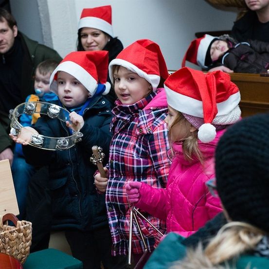 Eine Gruppe von Kindern mit Weihnachtsmützen spielt Musikinstrumente, darunter eine Schelle, in einer festlichen Umgebung. Hinter ihnen sitzen Erwachsene und Kinder, einige tragen Mützen, andere schlafen.