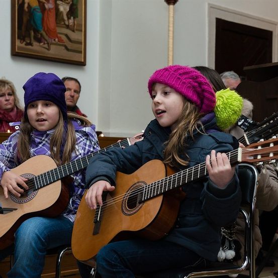 Zwei junge Mädchen sitzen auf Stühlen und spielen Gitarren, mit einem dritten Mädchen mit grüner Mütze hinter ihnen. Sie sind in einem Raum mit einem Gemälde an der Wand und einem Mikrofon auf einem Ständer.