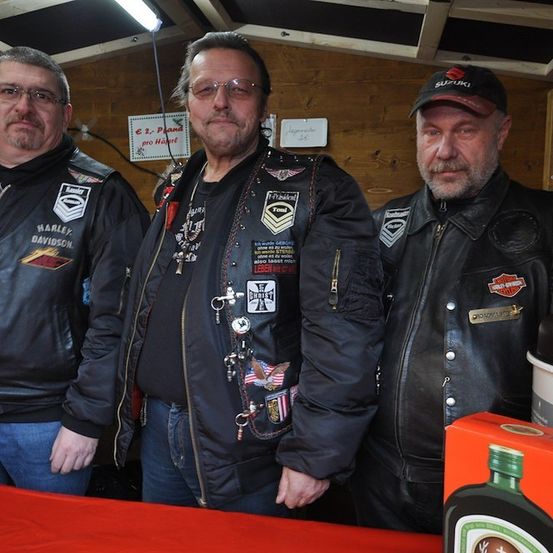 Drei Männer in Lederjacken stehen in einem Raum, einer trägt eine Kappe mit Logo, ein anderer ein Harley-Davidson-Patch und der dritte verschiedene Abzeichen. Eine Flasche und eine Schachtel stehen auf einem Tisch vor ihnen.