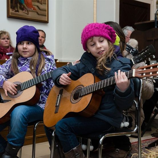 Zwei junge Mädchen spielen Gitarren in einem Raum, eines trägt einen rosa Hut und das andere einen lila Hut. Hinter ihnen sitzen einige Leute und schauen zu.