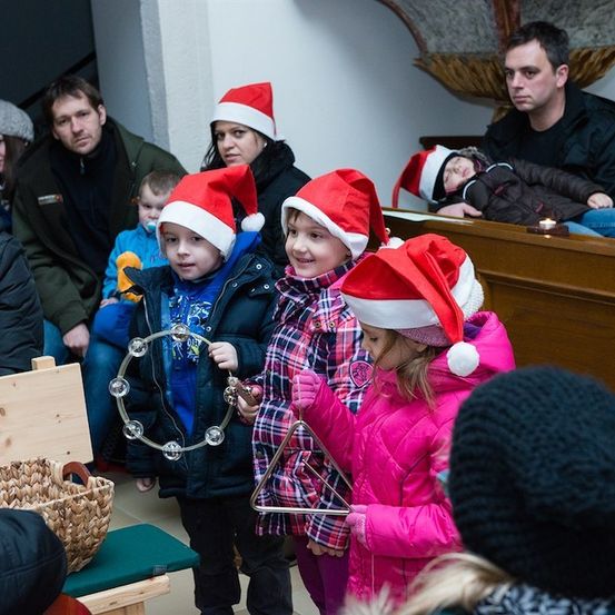 Eine Gruppe von Kindern in Weihnachtsmützen spielt Musikinstrumente in einem Raum, während Erwachsene zusehen.