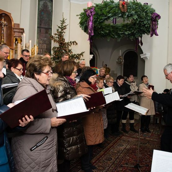 Ein Chor von Menschen in Winterkleidung singt in einer Kirche, angeführt von einem Dirigenten. Die Kirche ist mit Weihnachtskränzen und Kerzen geschmückt.