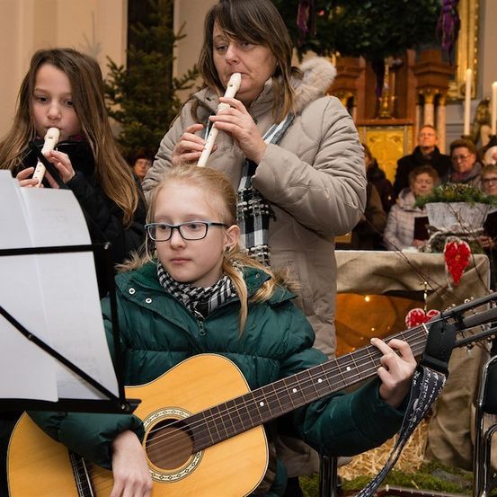 Drei Mädchen spielen Musikinstrumente in einer Kirche, wobei eines die Gitarre, ein anderes die Blockflöte und eine Frau hinter ihnen die Blockflöte spielt.