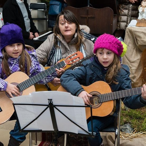 Drei junge Mädchen, zwei in lila und rosa Mützen, spielen Gitarren mit einer Frau in einer beigen Jacke. Sie sitzen auf Stühlen mit einem Notenständer vor ihnen. Dahinter steht ein Klavier und ein Tisch mit einer Statue.