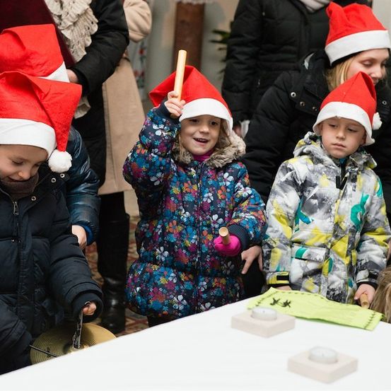 Mehrere Kinder mit Weihnachtsmützen versammeln sich um einen Tisch und halten Trommelstöcke und Becken in der Hand, wahrscheinlich an einer festlichen Aktivität teilnehmend.