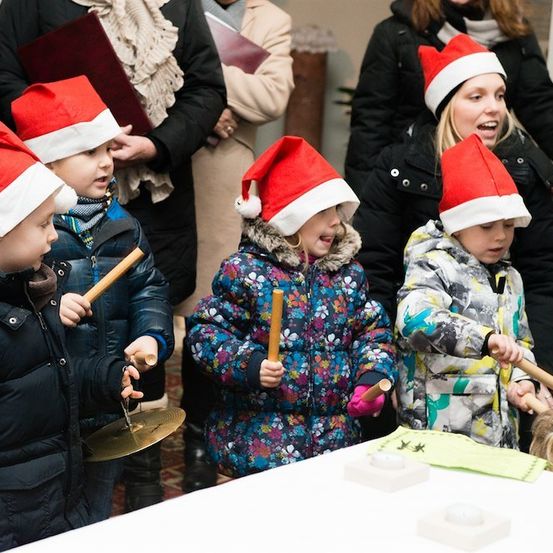 Fünf Kinder in Weihnachtsmützen spielen Instrumente, darunter Becken und Trommelstöcke, in einer festlichen Umgebung. Dahinter beobachten Erwachsene, einer von ihnen lächelt.