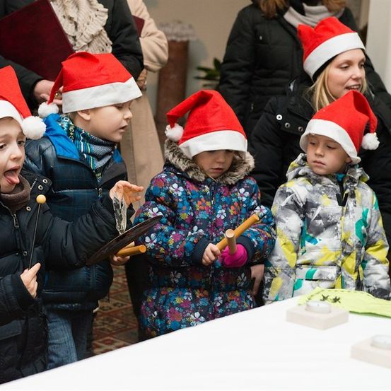 Eine Gruppe von Kindern mit Weihnachtsmützen spielt Musikinstrumente in einer festlichen Umgebung.