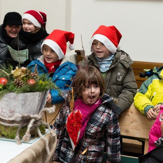 Eine Gruppe von Kindern in Winterkleidung und Weihnachtsmützen versammelt sich um eine dekorierte Pflanze, möglicherweise für eine Weihnachtsveranstaltung.