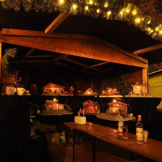 Ein hölzerner Pavillon mit einer weihnachtlichen Krippenaufstellung, beleuchtet von warmen Lichtern, mit einer Flasche und Gläsern auf einem Tisch davor.
