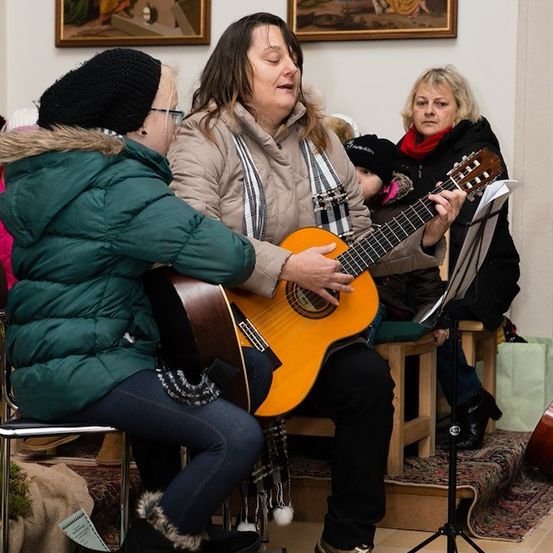 Eine Frau spielt Gitarre, während eine andere Frau zusieht, beide sitzen in einem Raum mit weißer Wand und zwei Gemälden.