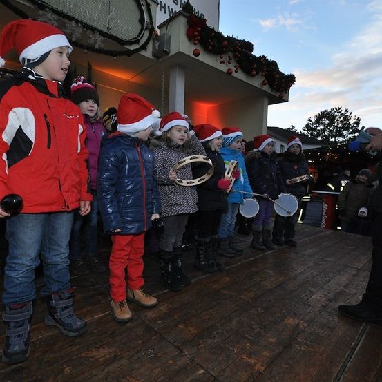 Eine Gruppe von Kindern in Winterkleidung und Weihnachtsmützen führt eine musikalische Darbietung auf einer hölzernen Bühne auf. Ein Mann steht neben ihnen, möglicherweise ihr Lehrer. Die Bühne ist mit Weihnachtsdekorationen geschmückt.