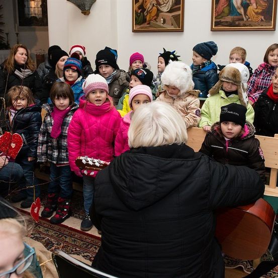 Eine Gruppe von Kindern in Winterkleidung versammelt sich um eine Frau, die eine Gitarre spielt, mit Erwachsenen und weiteren Kindern im Hintergrund.
