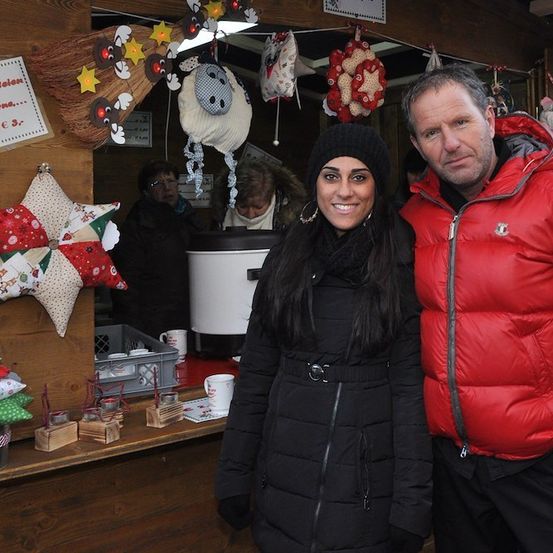 Ein Mann und eine Frau stehen lächelnd vor einem festlich geschmückten Weihnachtsmarktstand und posieren für die Kamera. Der Stand ist mit Weihnachtsdekorationen und einem Schild, das 'Weihnachtsgeschenke' besagt, geschmückt.