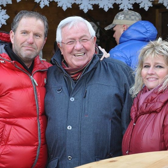 Drei Menschen stehen eng beieinander und lächeln für ein Foto. Der Mann links trägt einen roten Mantel, der Mann in der Mitte einen blauen Mantel und die Frau rechts einen roten Mantel. Hinter ihnen ist ein Mann in einem blauen Mantel und einer Mütze zu sehen.