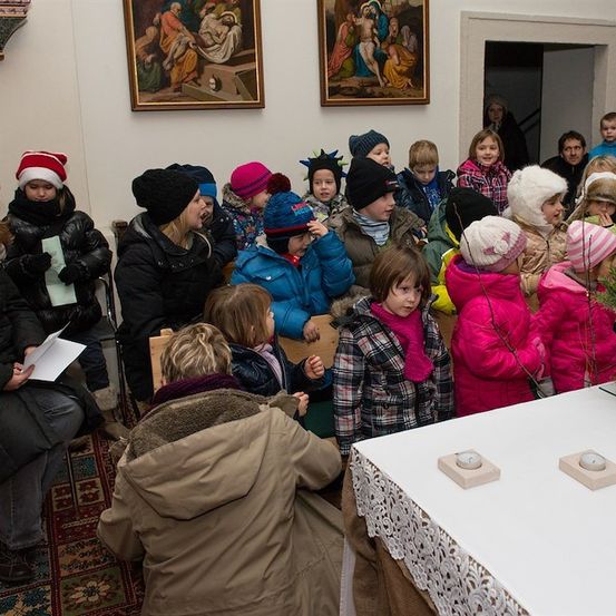 Eine Gruppe von Kindern und Erwachsenen ist drinnen versammelt, möglicherweise in einer Kirche, und trägt Winterkleidung. Einige sitzen, andere stehen, und ein Tisch mit Kerzen ist im Vordergrund.