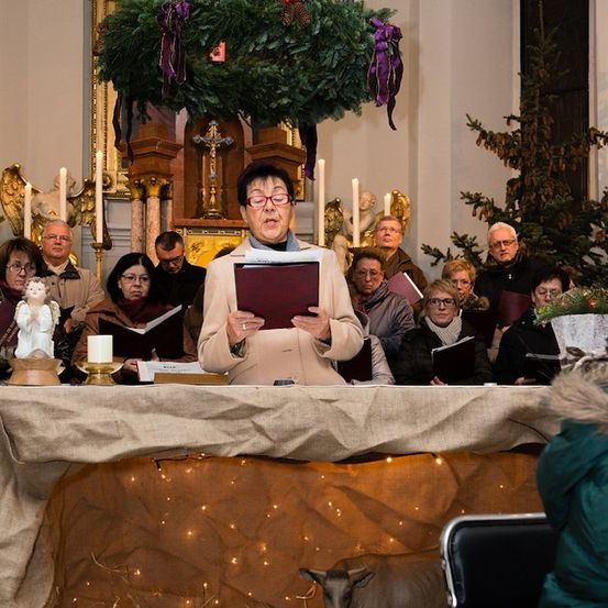 Ein Chor in Winterkleidung singt in einer für Weihnachten geschmückten Kirche. Eine Frau steht vorne und hält ein Notenbuch.