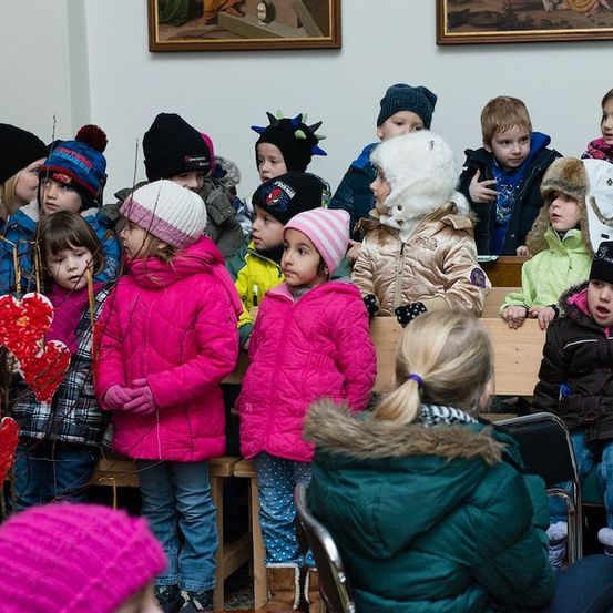 Eine Gruppe von Kindern in Winterkleidung steht in einem Raum, einige halten herzförmige Dekorationen. Eine erwachsene Frau mit einem Pferdeschwanz sitzt vor ihnen.