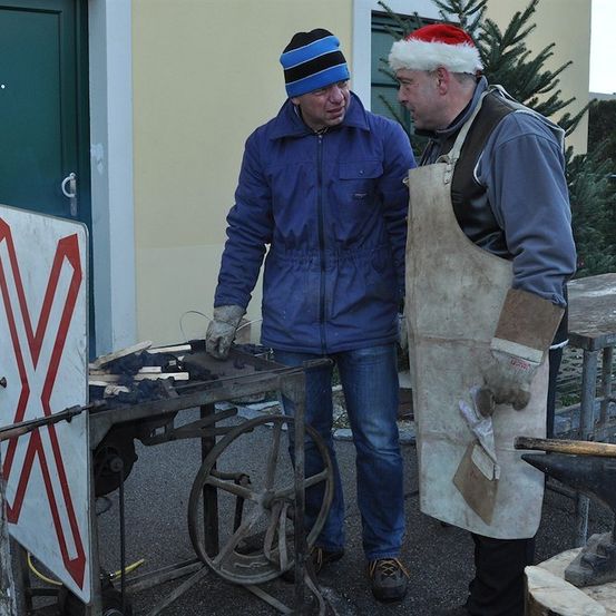 Zwei Männer stehen vor einem Metalltisch mit einem roten X-Zeichen, einer trägt einen Weihnachtsmannhut und eine Schürze, der andere eine Jacke und Handschuhe. Sie machen wahrscheinlich etwas Arbeit. Hinter ihnen steht ein Weihnachtsbaum.